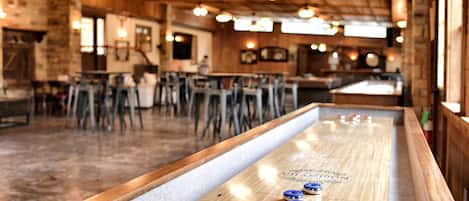 A bar area with a shuffleboard table, wooden bar stools, and hanging lights.