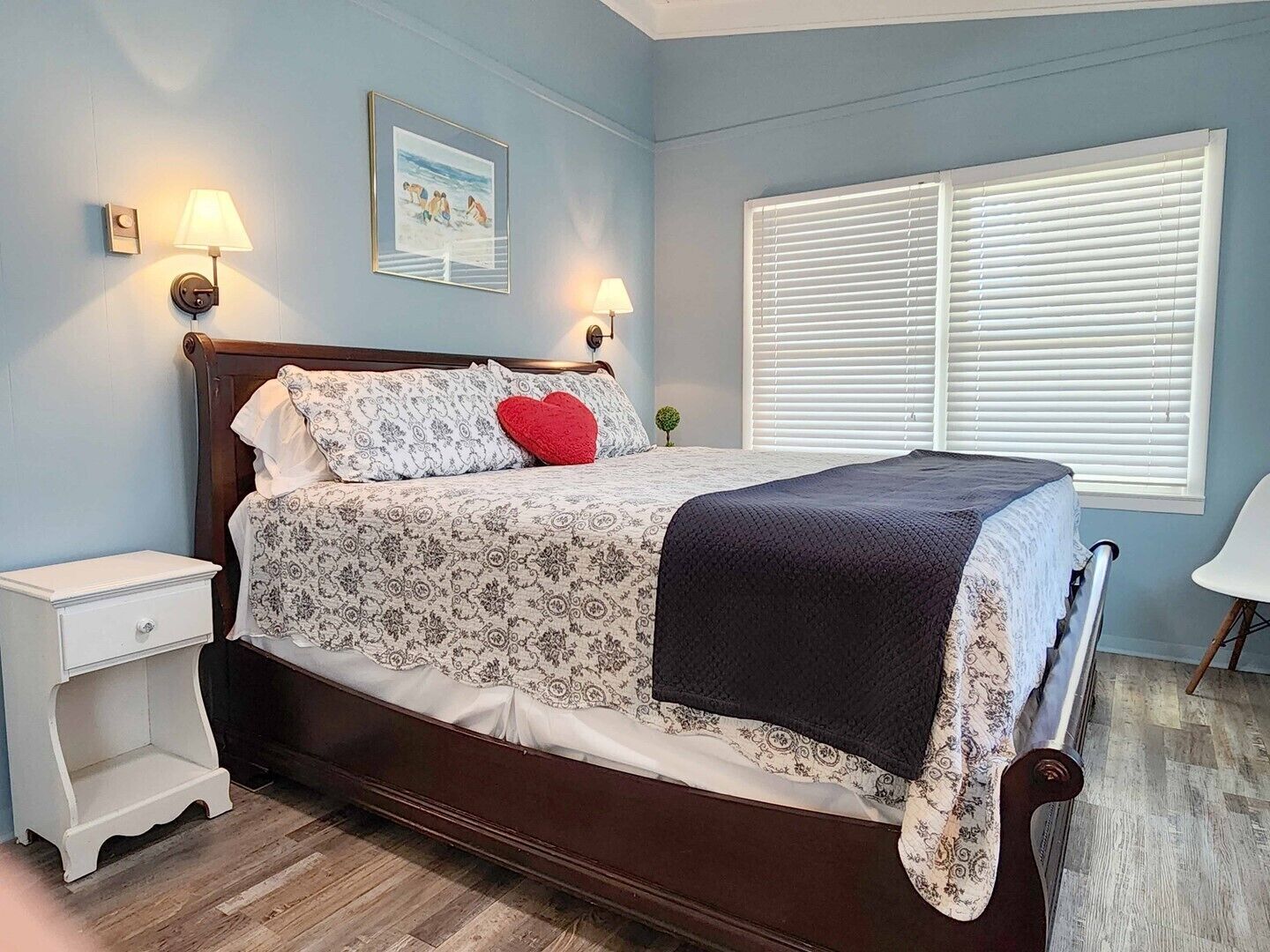 A bedroom with a wooden bed, two bedside lamps, a window with blinds, and a small bedside table.