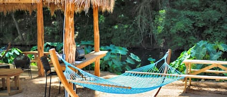 A hammock, wooden chairs, a table, and a thatched-roof structure in a garden setting.