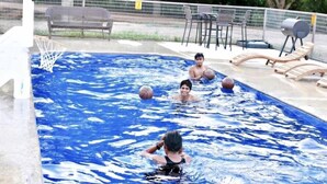 Una piscina amb un cèrcol de bàsquet, seients de relax i una zona enclotada amb arbres.