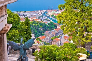 Une vue urbaine côtière avec une statue de dragon au premier plan, un port avec des bateaux, et un mélange de bâtiments historiques et modernes.
