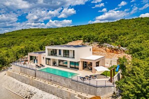 Une villa moderne dotée d’une piscine, d’un espace salon extérieur et offrant une vue sur une colline boisée.
