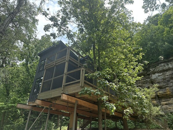 Ein Baumhaus mit einer Holzdachterrasse und einem Metallgeländer, umgeben von dichtem Wald.