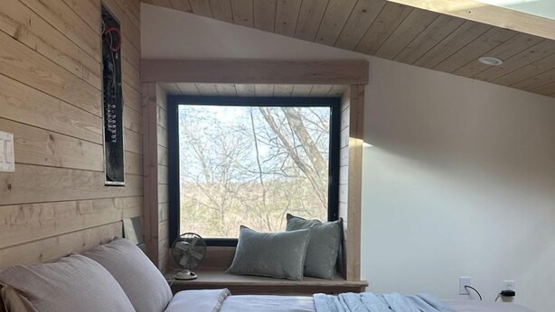 Un dormitorio con techo de madera, un ventanal grande, una cama con almohadas y una mesita de noche.