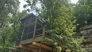 A treehouse with a wooden deck and a metal railing, surrounded by dense forest.