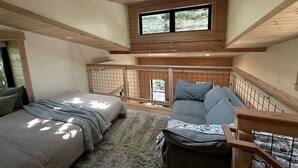 A bedroom with two beds, a sofa, a wooden ceiling, and a window overlooking greenery.