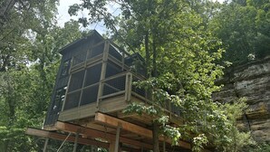 Una casa en los árboles con terraza de madera y barandilla de metal, rodeada de un bosque frondoso.