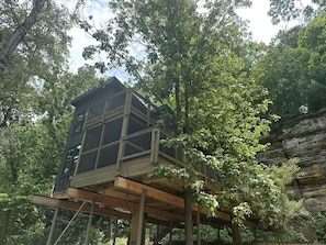 Una casa en los árboles con terraza de madera y barandilla de metal, rodeada de un bosque frondoso.