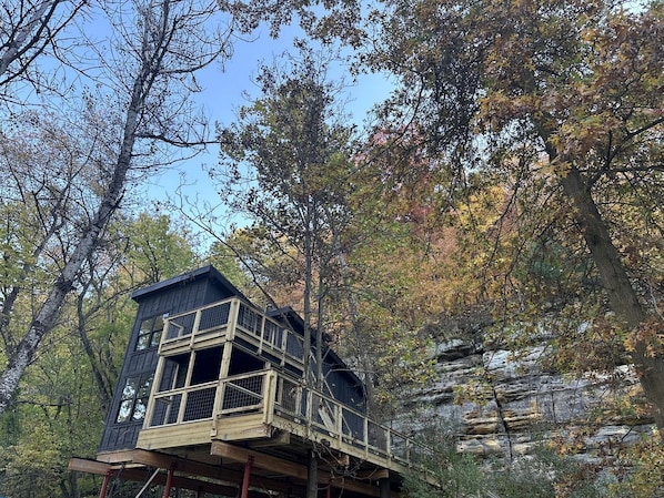 A wooden cabin with a balcony, surrounded by trees and a rocky cliff.