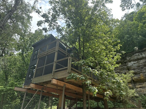 A treehouse with a wooden deck and a metal railing, surrounded by dense forest.