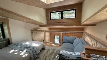 A bedroom with two beds, a sofa, a wooden ceiling, and a window overlooking greenery.