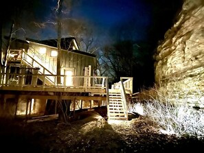 Eine Holzhütte mit einer Treppe, die zu einer Terrasse führt, umgeben von Bäumen und einer Felsformation.