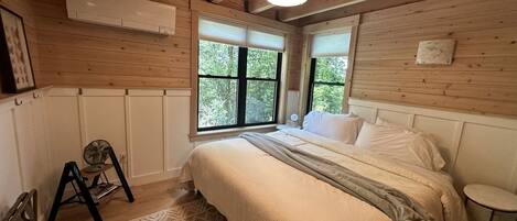 A bedroom with a bed, a standing fan, a round table, and a window with a view of trees.