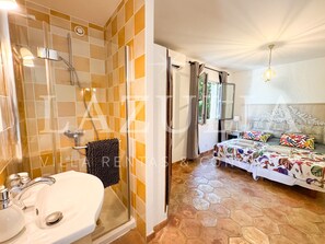 A bathroom with a glass shower enclosure, a sink, and a toilet. The bedroom features a bed with a floral bedspread, a nightstand, and a window with curtains. - Villa Boucheron - 4 independent apartments - Sea view (Roquebrune-sur-Argens)