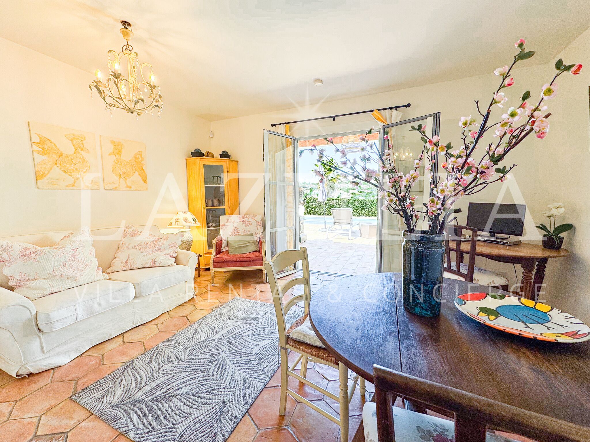 Un salon comprenant un canapé, une table à manger avec des chaises, un lustre et une vue sur l’extérieur, visible à travers une porte vitrée.