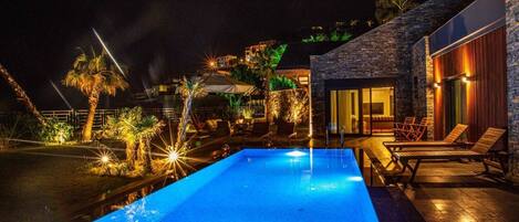 A nighttime swimming pool with lounge chairs and a stone building with a glass door.