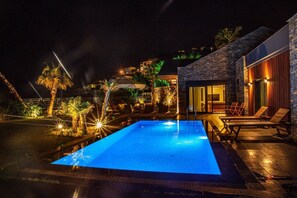 Ein Pool bei Nacht mit Liegestühlen und einem Steinhaus mit Glastür.