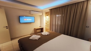 A hotel room with a bed, a TV mounted on the wall, an air conditioning unit, a small table, and a curtain. - Elisa Luxury Suite in Nea Flogita (Flogita)