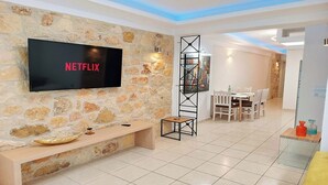 Ein Wohnzimmer mit Steinwand, einem Flachbildfernseher, einer hölzernen Bank, einem hohen, schwarzen Metallregal und einem Essbereich mit weißen Stühlen und einem Tisch.
