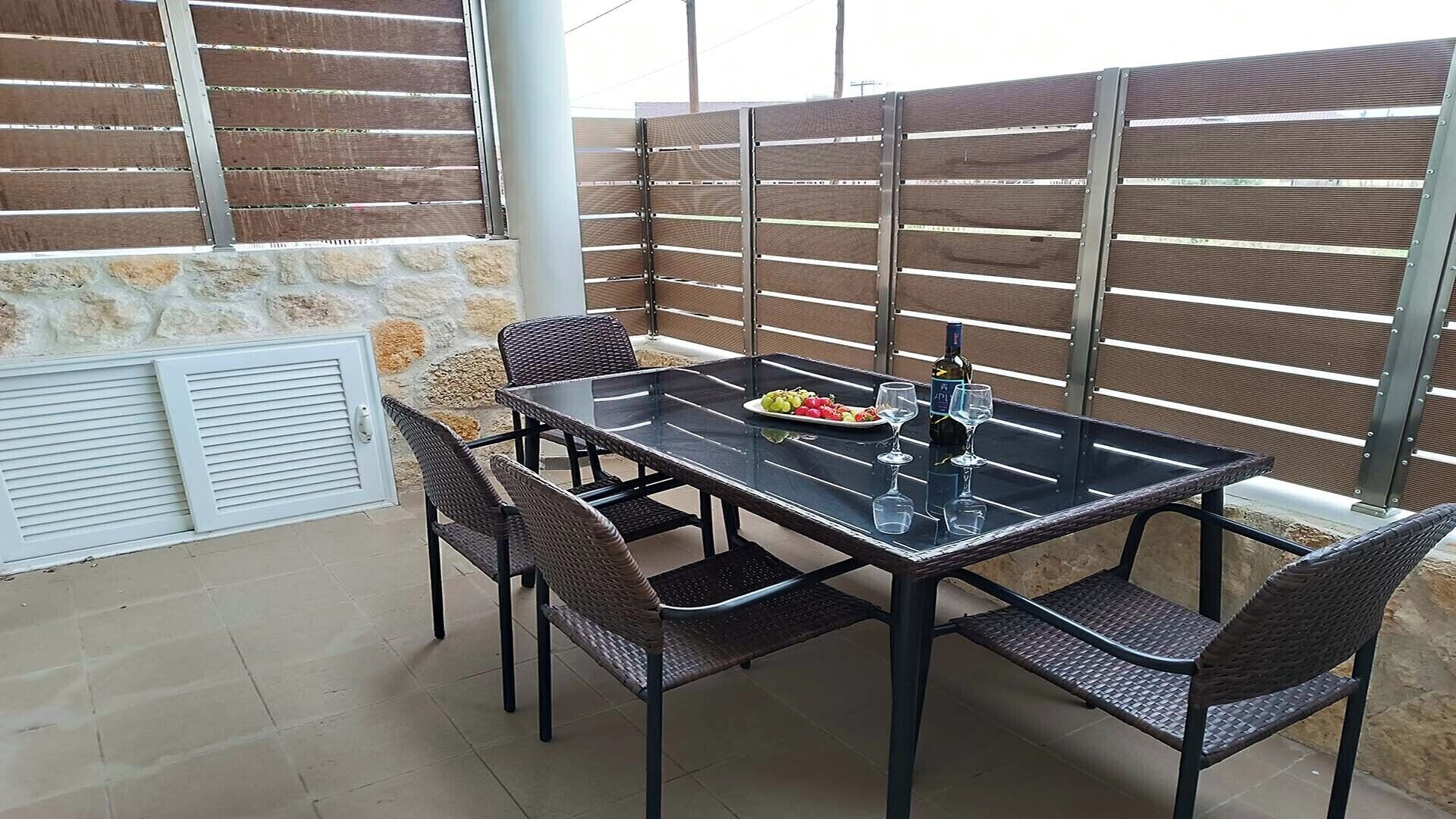 An outdoor patio with a glass-top table, four wicker chairs, a bottle of wine, and a plate of fruit.