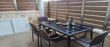 An outdoor patio with a glass-top table, four wicker chairs, a bottle of wine, and a plate of fruit.