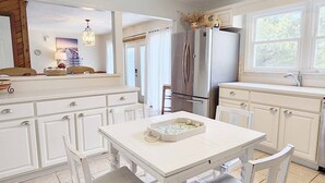 A kitchen with white cabinetry, a stainless steel refrigerator, a dining table with chairs, and a window with curtains. - 600 Steps, Great Family Beach House, Pet friendly & Close to Beach (Pawleys Island)