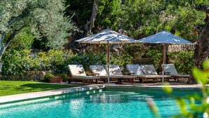 Ein Pool mit klarem Wasser, umgeben von Liegestühlen und Sonnenschirmen, mit Bäumen und einer Steinmauer im Hintergrund.