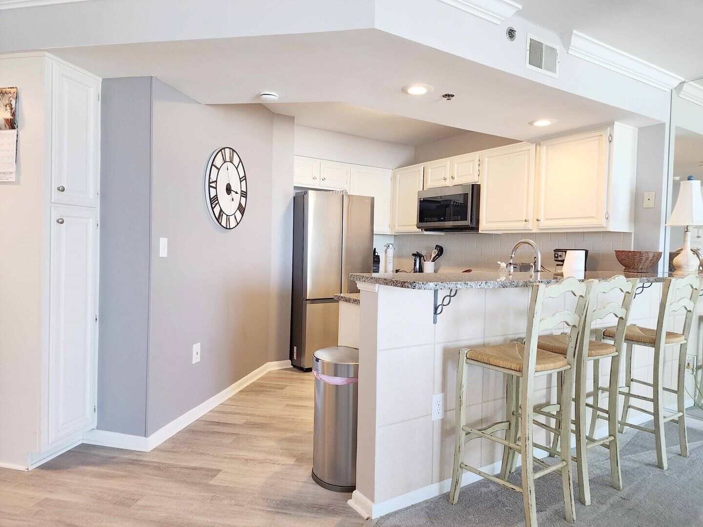 A modern kitchen with white cabinets, a stainless steel refrigerator, a microwave, and a bar area with stools.