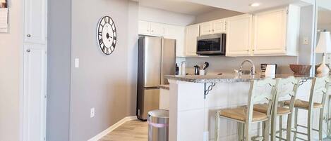 A modern kitchen with white cabinets, a stainless steel refrigerator, a microwave, and a bar area with stools.