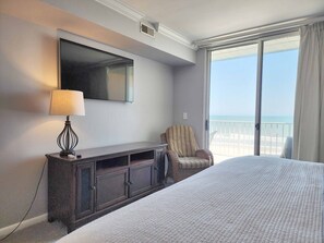 A hotel room with a bed, a TV, a desk with a lamp, a chair, and a balcony with a view of the beach. - Hamilton 502- Direct Ocean View from Top Floor (Pawleys Island)