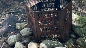 A metal fire pit with the words 'CAMPING CAN'T STOP WONDER' engraved on it, surrounded by rocks and a picnic table in a wooded area.