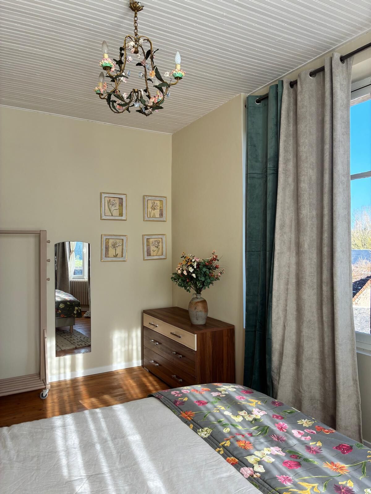 Une chambre à coucher comprenant un lit, une commode, un miroir, des œuvres d’art florales et un lustre.