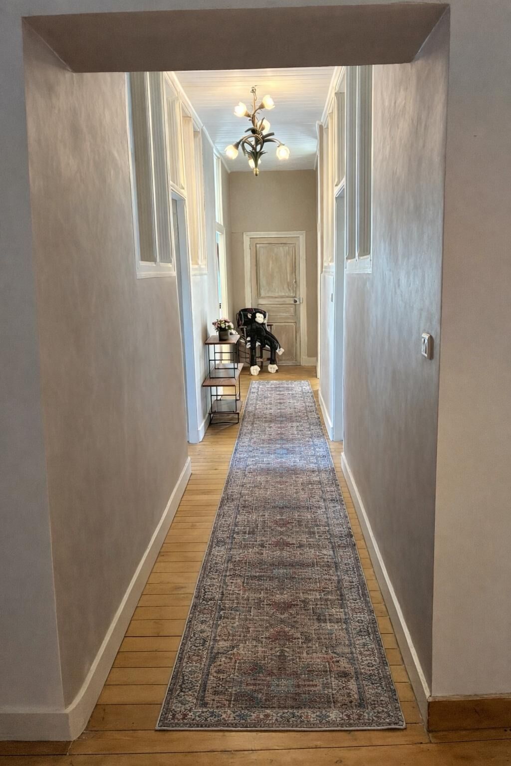 A hallway with a patterned rug, a chandelier, and a door at the end.