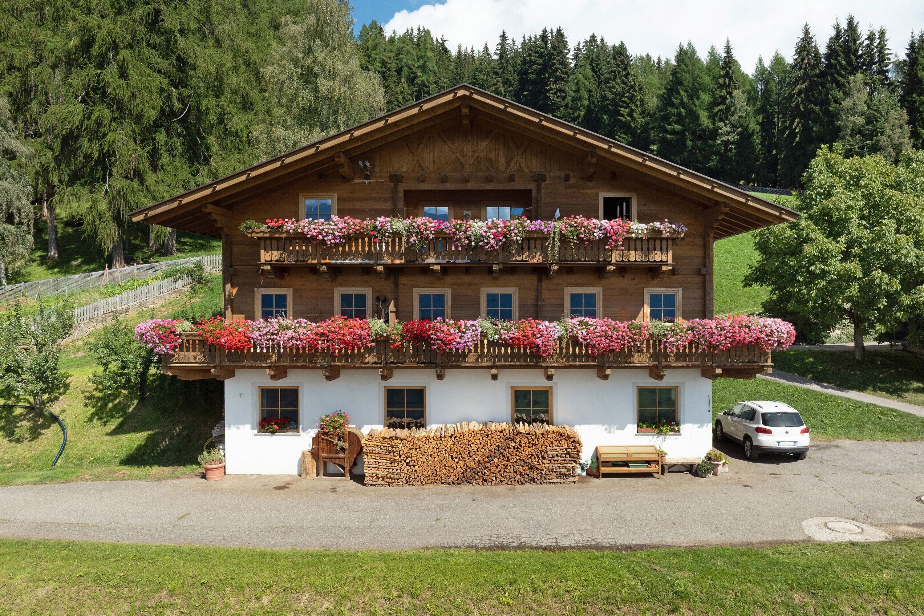 Un chalet en bois à deux étages, doté de balcons ornés de fleurs, d’un banc en bois et d’une voiture garée devant.