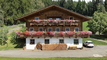 Un chalet en bois à deux étages, doté de balcons ornés de fleurs, d’un banc en bois et d’une voiture garée devant.