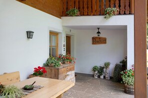 Une table en bois avec un pot de fleurs, un banc en bois et une pancarte indiquant « Penel-1-boy », dans une cour dallée de pierres et agrémentée de plantes en pot.