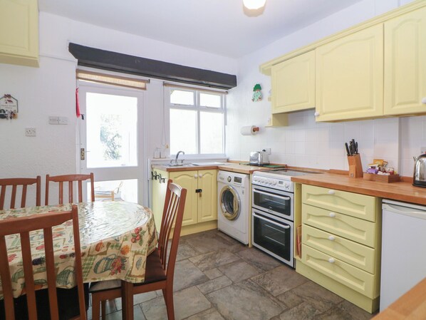A compact kitchen with a washing machine, oven, and a dining table with chairs. - Honeysuckle Cottage (Buxton)