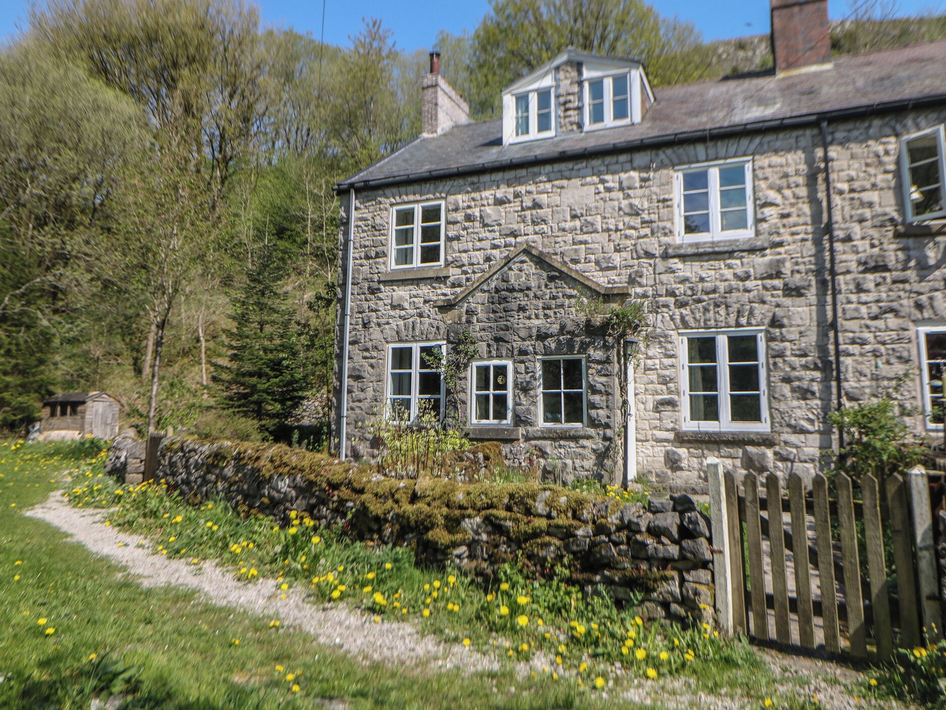 A stone house with a gabled roof, a small attached shed, and a stone wall with yellow flowers.