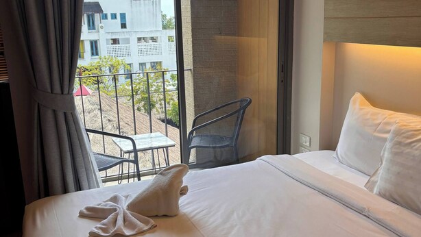 A hotel room with a bed, a chair, a table, and a view of buildings outside. - Coconut Studio Aonang (Krabi)