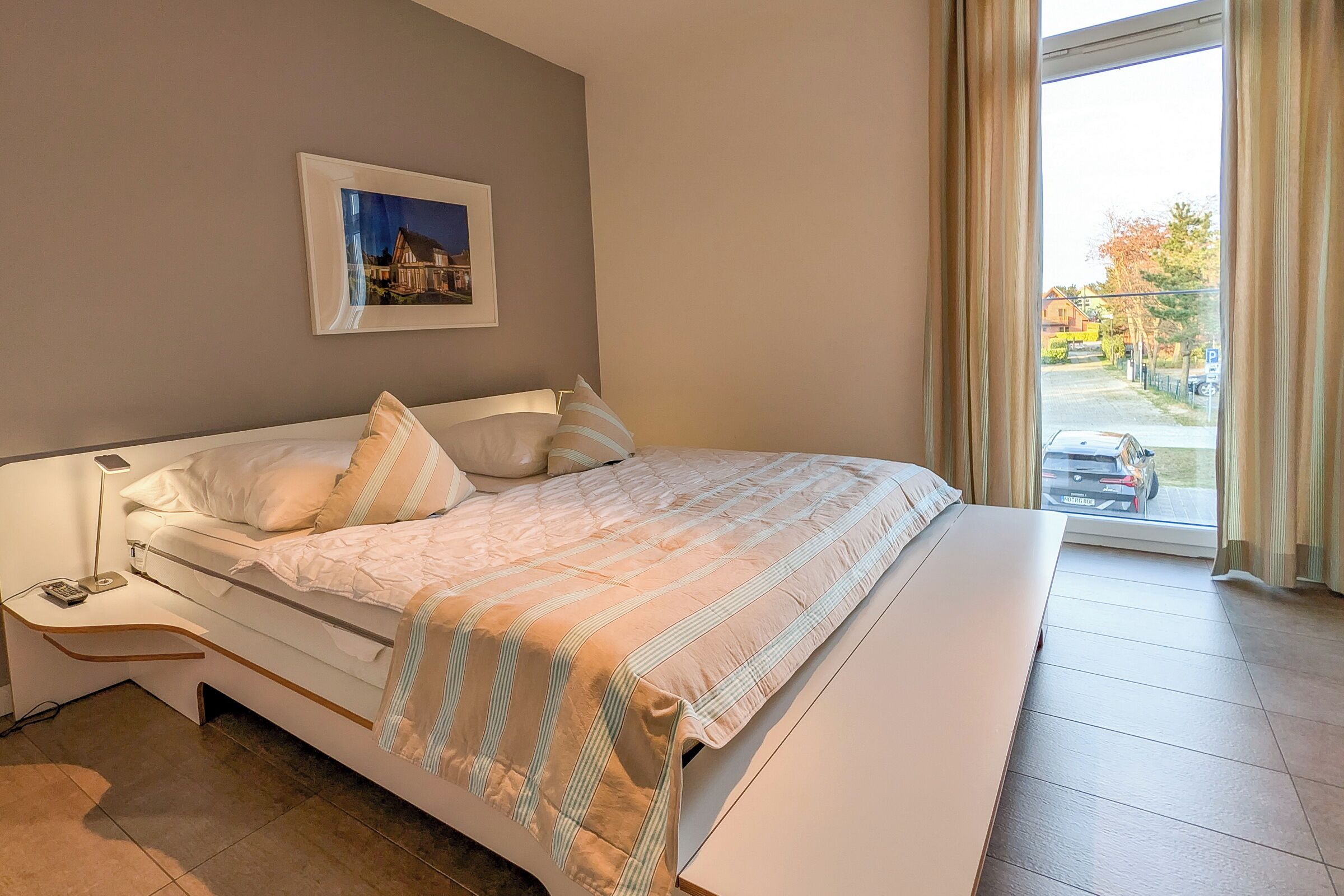 A bedroom with a bed, bedside table, lamp, and a window with a view of the outside.