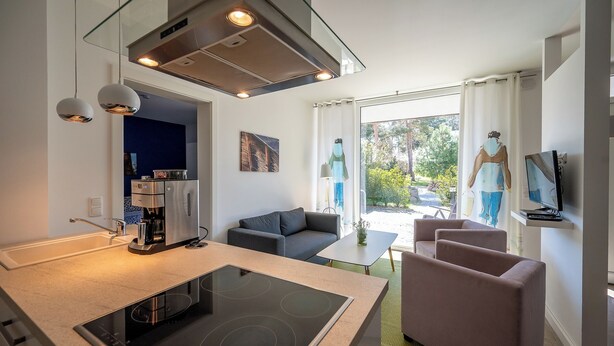 Eine moderne Küche mit Einbauherd, Kaffeemaschine und Pendelleuchten. Im Wohnbereich befindet sich eine Couch und ein Fernseher, mit Blick ins Freie.