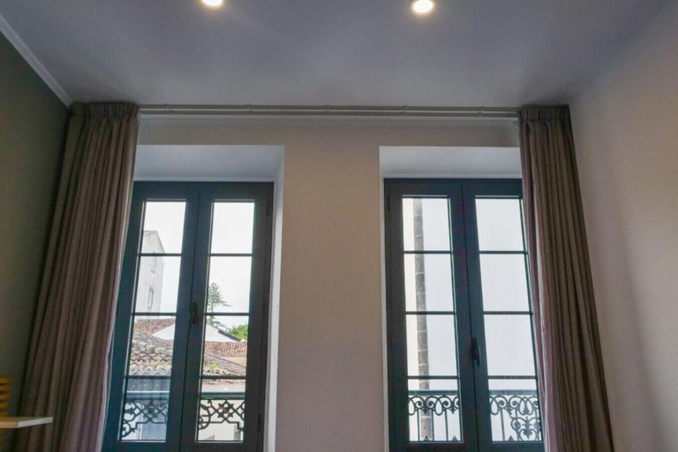 A room with two large windows, a balcony with a metal railing, and a ceiling with recessed lighting.