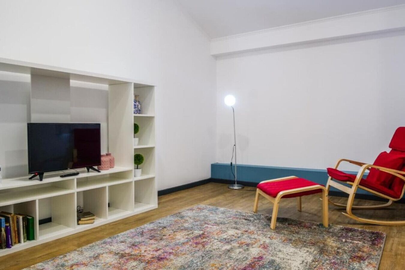 Un salon avec un téléviseur, une bibliothèque, un fauteuil rouge, une lampe sur pied et un tapis à motifs.
