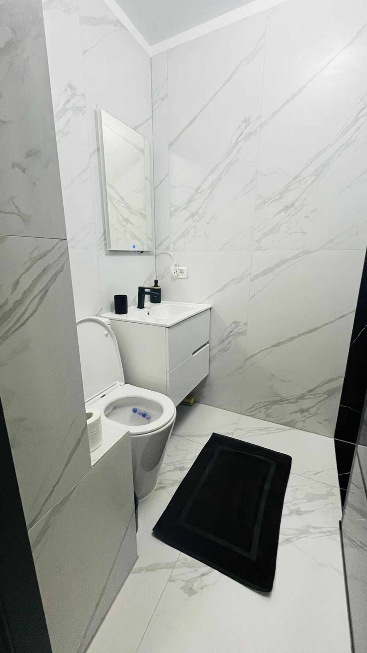 A bathroom with a toilet, sink, mirror, and black mat on the floor.