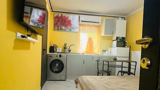 A compact room with a bed, a washing machine, a small table, and a television mounted on the wall.