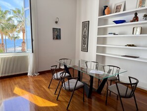 A dining area with a glass table and chairs, a white wall with artwork, a shelf with decorative items, and a view of the sea through a window. - Casa Catalina (Imperia)