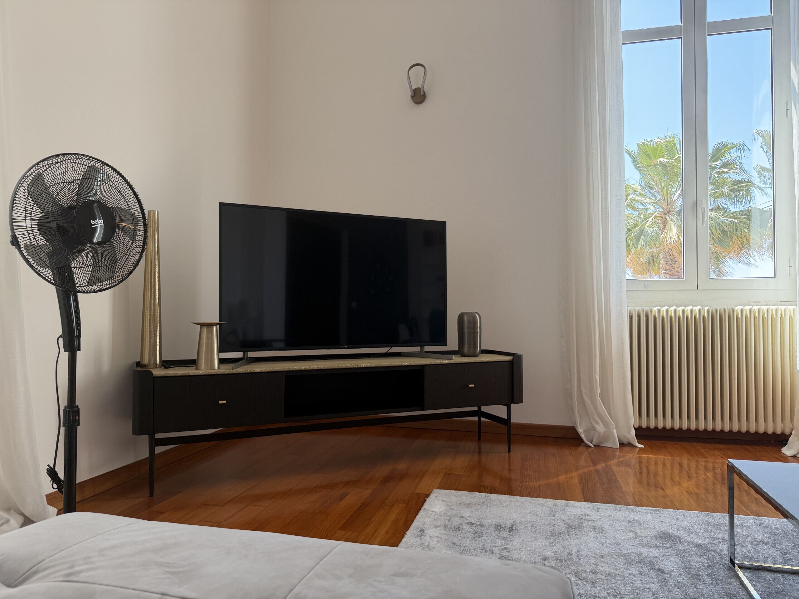 A modern living room with a flat-screen TV on a black TV stand, a standing fan, a radiator, and a large window with a view of palm trees.