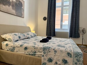 A bedroom with a bed, a clock, a window with curtains, and a standing fan. - Casa Catalina (Imperia)