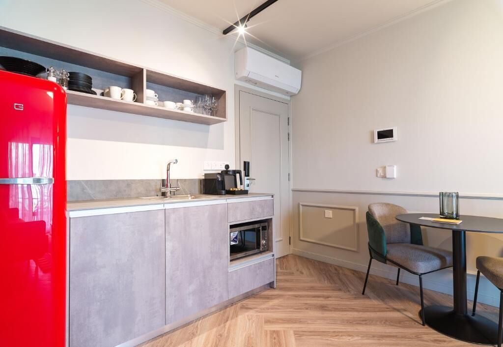 A compact kitchen with a red refrigerator, microwave, and oven, a dining area with a round table and chairs, and a built-in shelf above the counter.