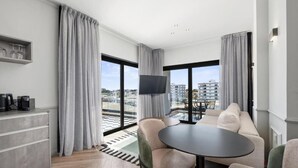 A modern hotel room with a sofa, a round table, a flat-screen TV, and a view of the cityscape through large windows.